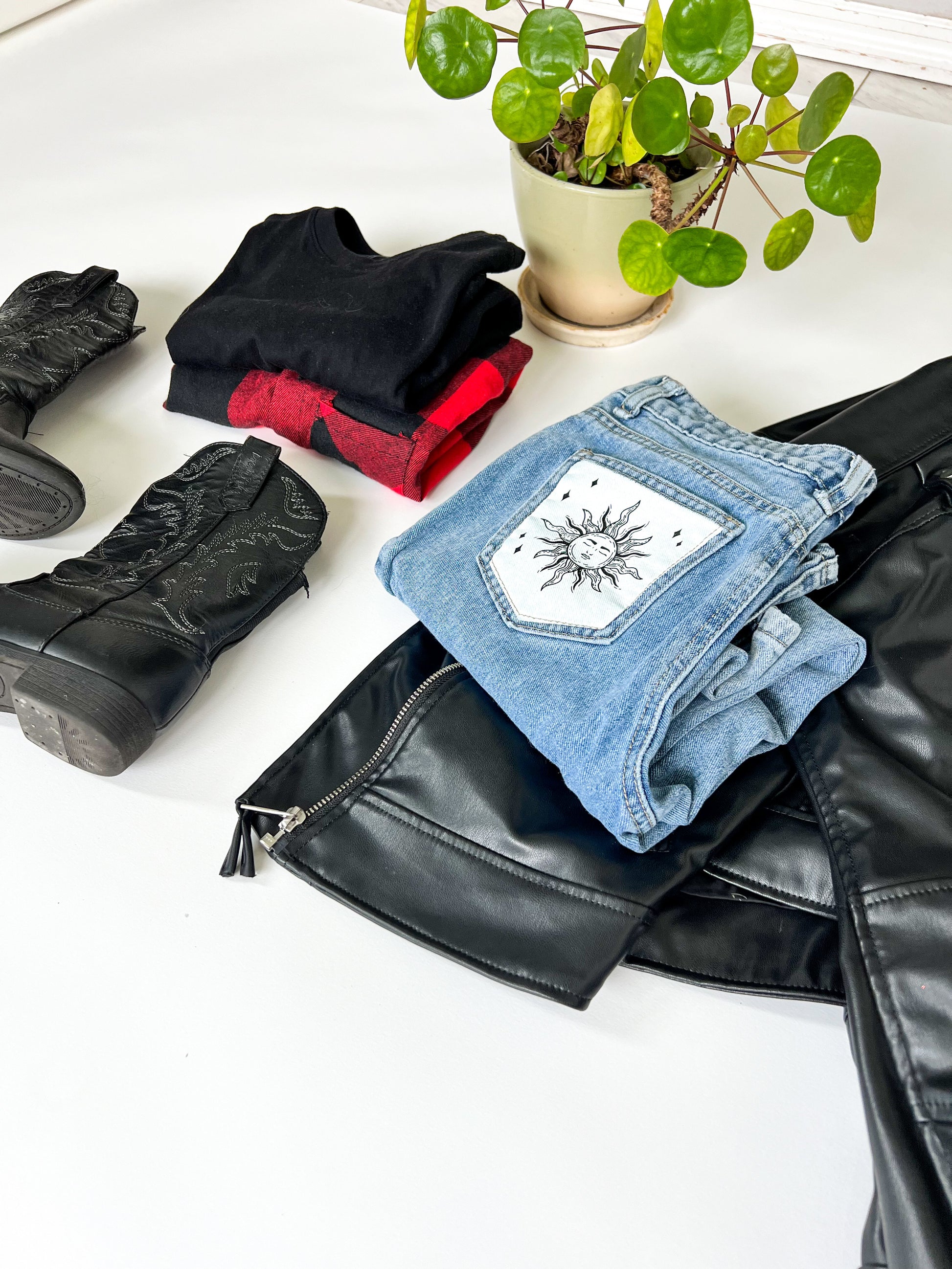Folded kids secondhand jeans with a sun design, black boots, and a plant on a white surface.